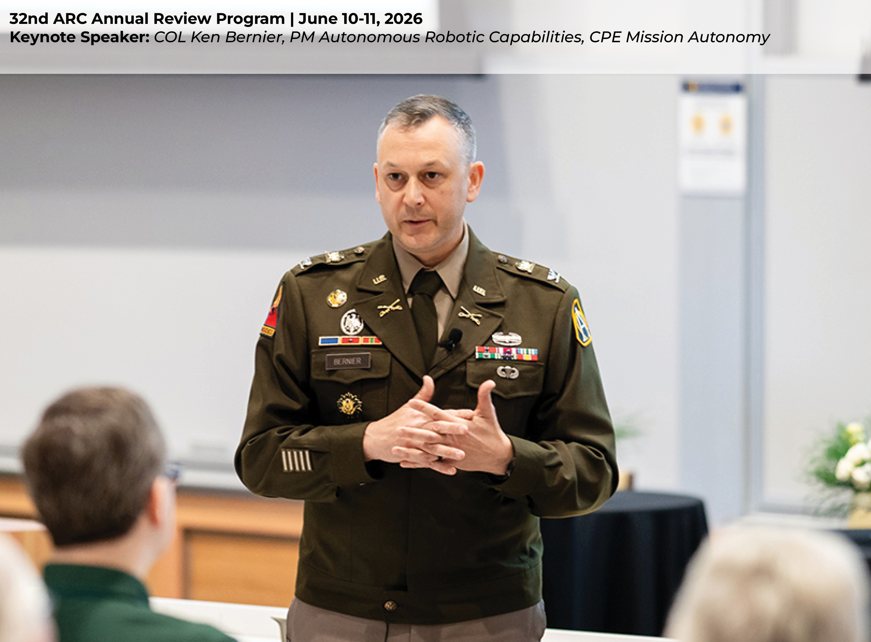 COL Ken Bernier giving a keynote presentation at a podium in an auditorium, addressing a seated audience during a formal event.