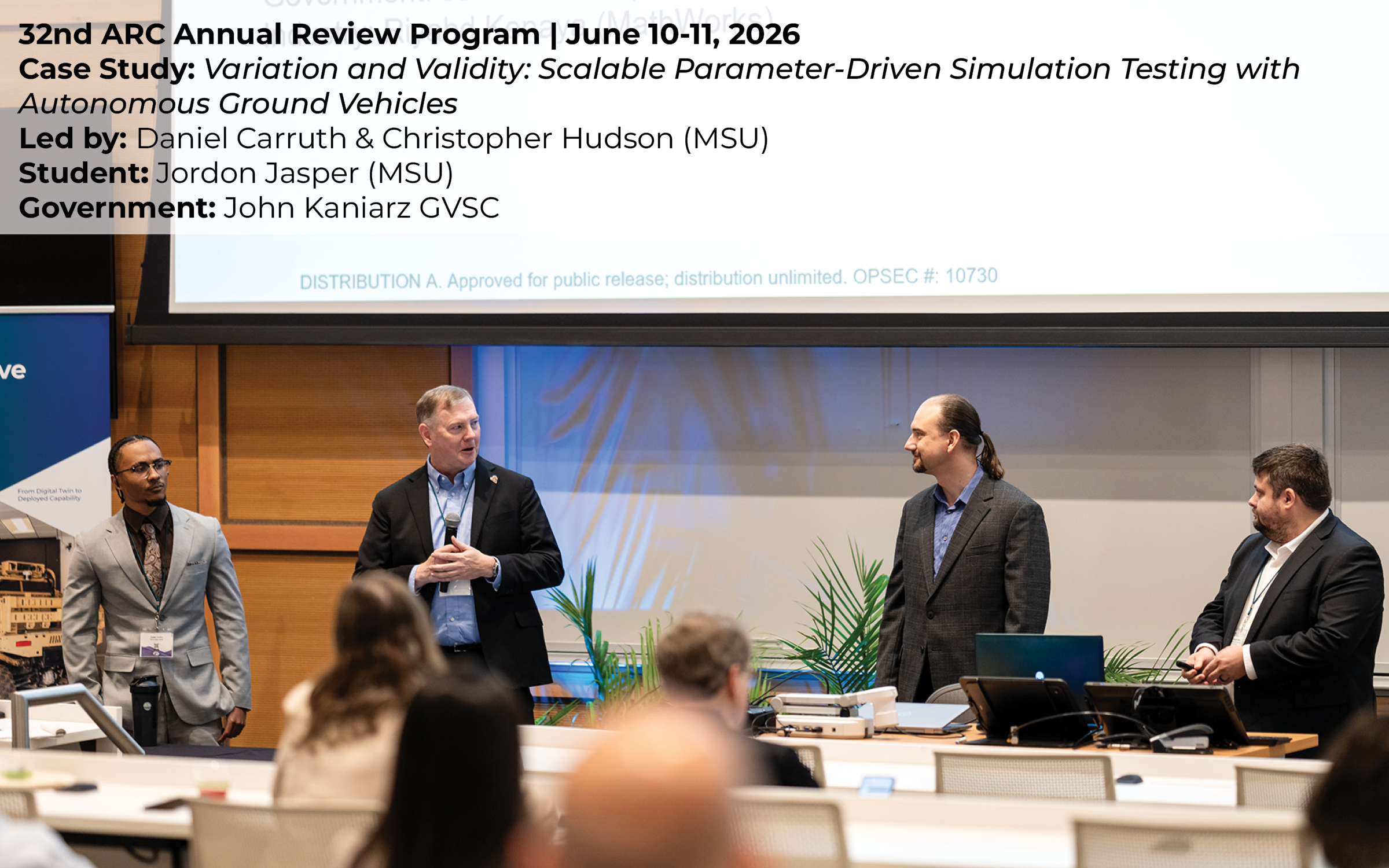 Four presenters standing at the front of an auditorium, addressing a seated audience during a formal presentation.