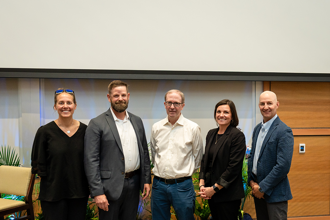 Kira Barton stands with panelists COL (Ret.) Warren Sponsler, Todd Allen, Jillyn Alban, and Scott James, for a group photo.