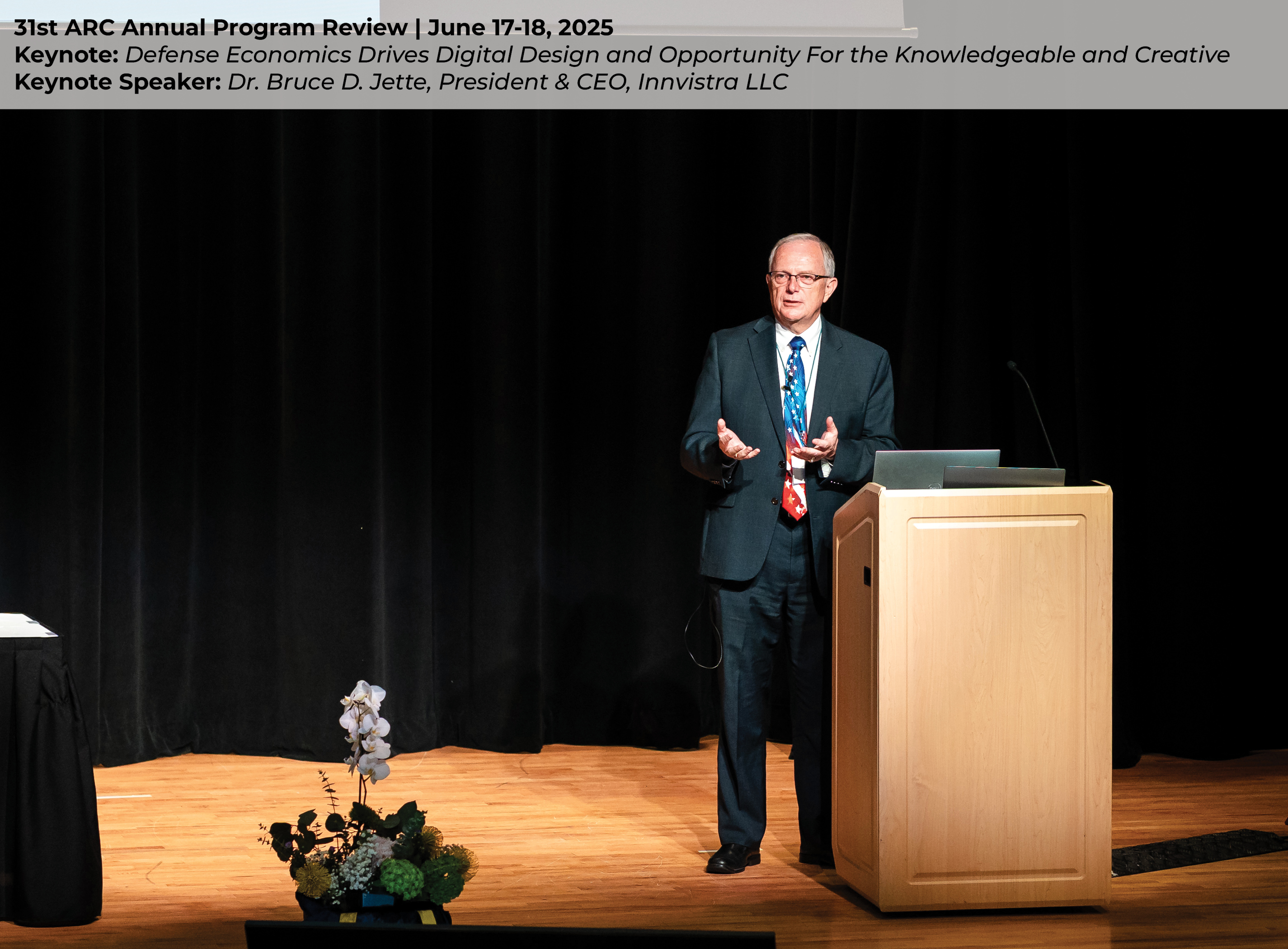 Dr. Bruce D. Jette on stage standing in front of a black curtain and behind a podium. He is delivering his keynote presentation “Defense Economics Drives Digital Design and Opportunity For the Knowledgeable and Creative”