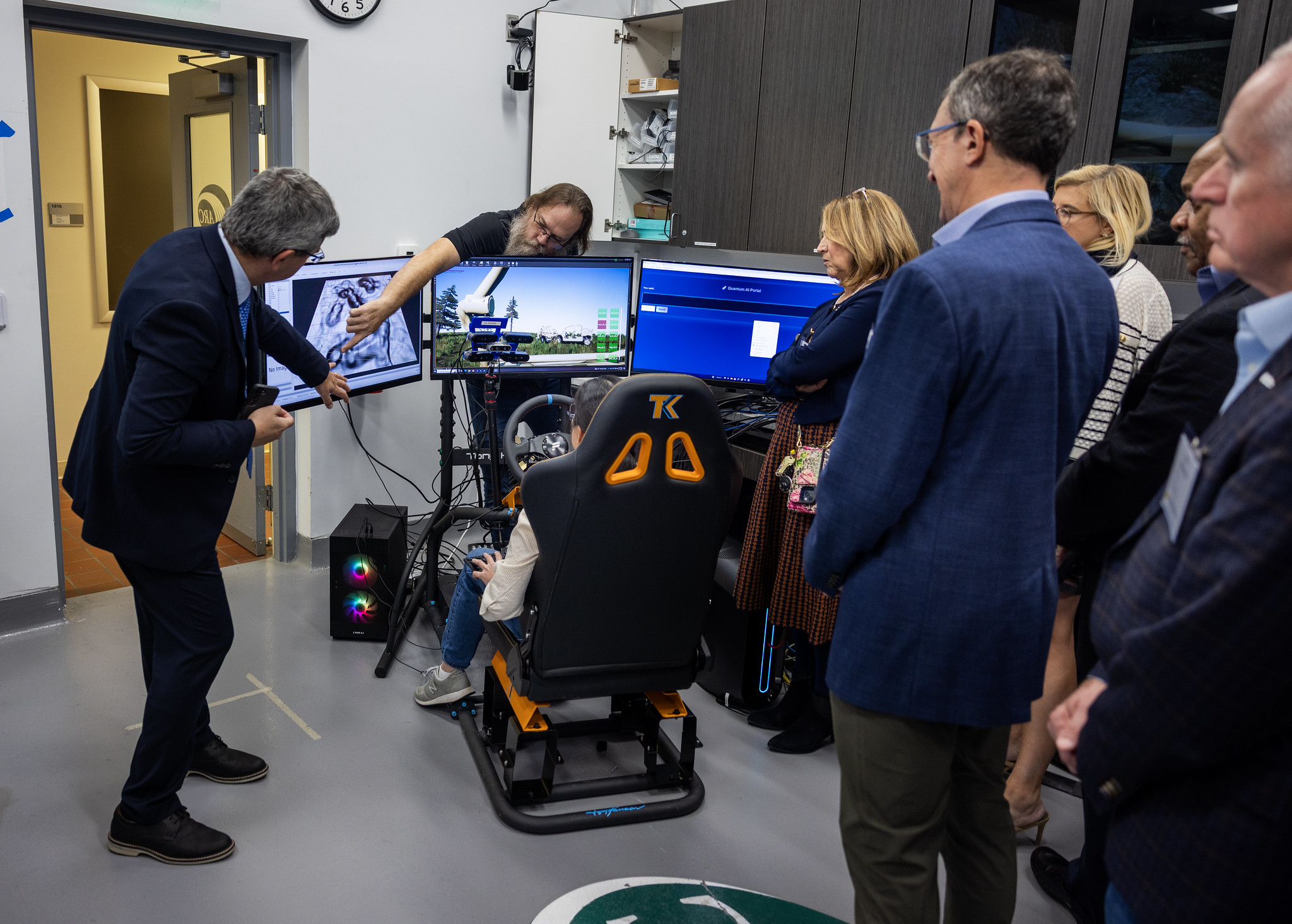 Prof. Bogdan Epureanu and Engineer Sean Rice standing behind three screens explaining the contents on the screen while participant sits in the black  demo seat 