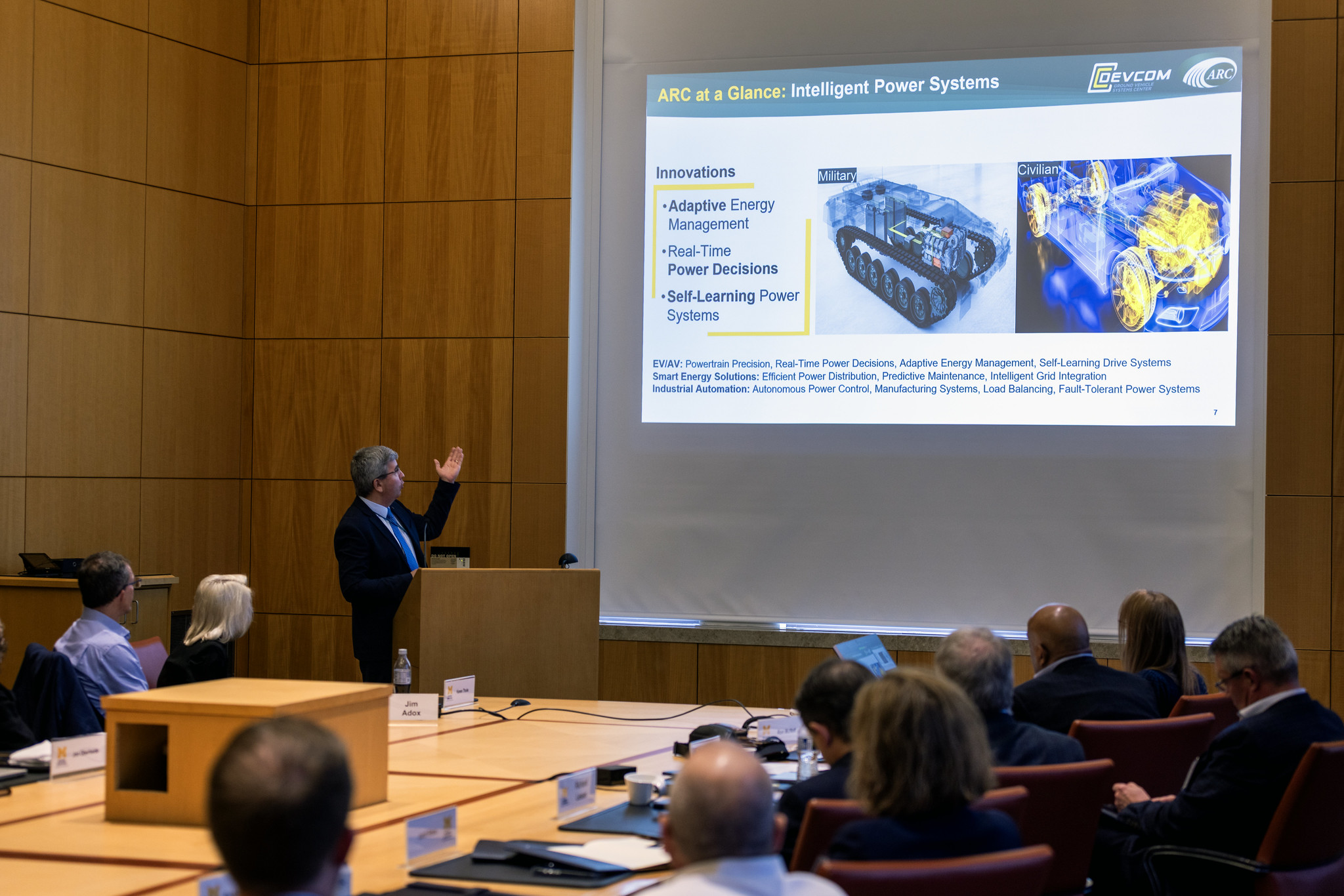 Prof. Bogdan Epureanu standing in front of a presentation screen and behind a podium, explaining the contents to members of an audience in board room.