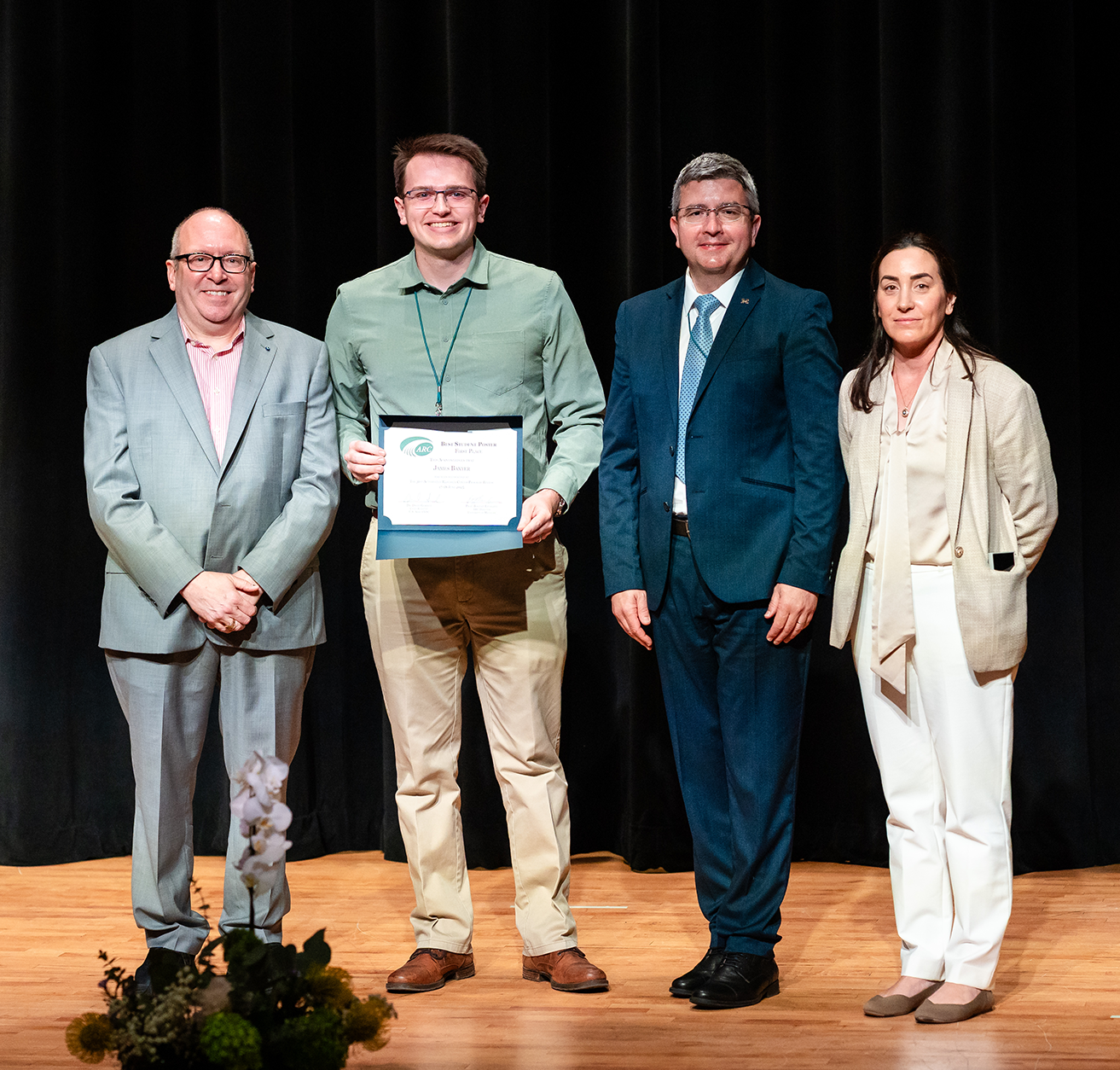 On stage from left to right: Dr. David Gorsich, James Baxter, Prof. Bogdan Epureanu, and Dr. Talia Sebastian, standing in front of a black curtain.
