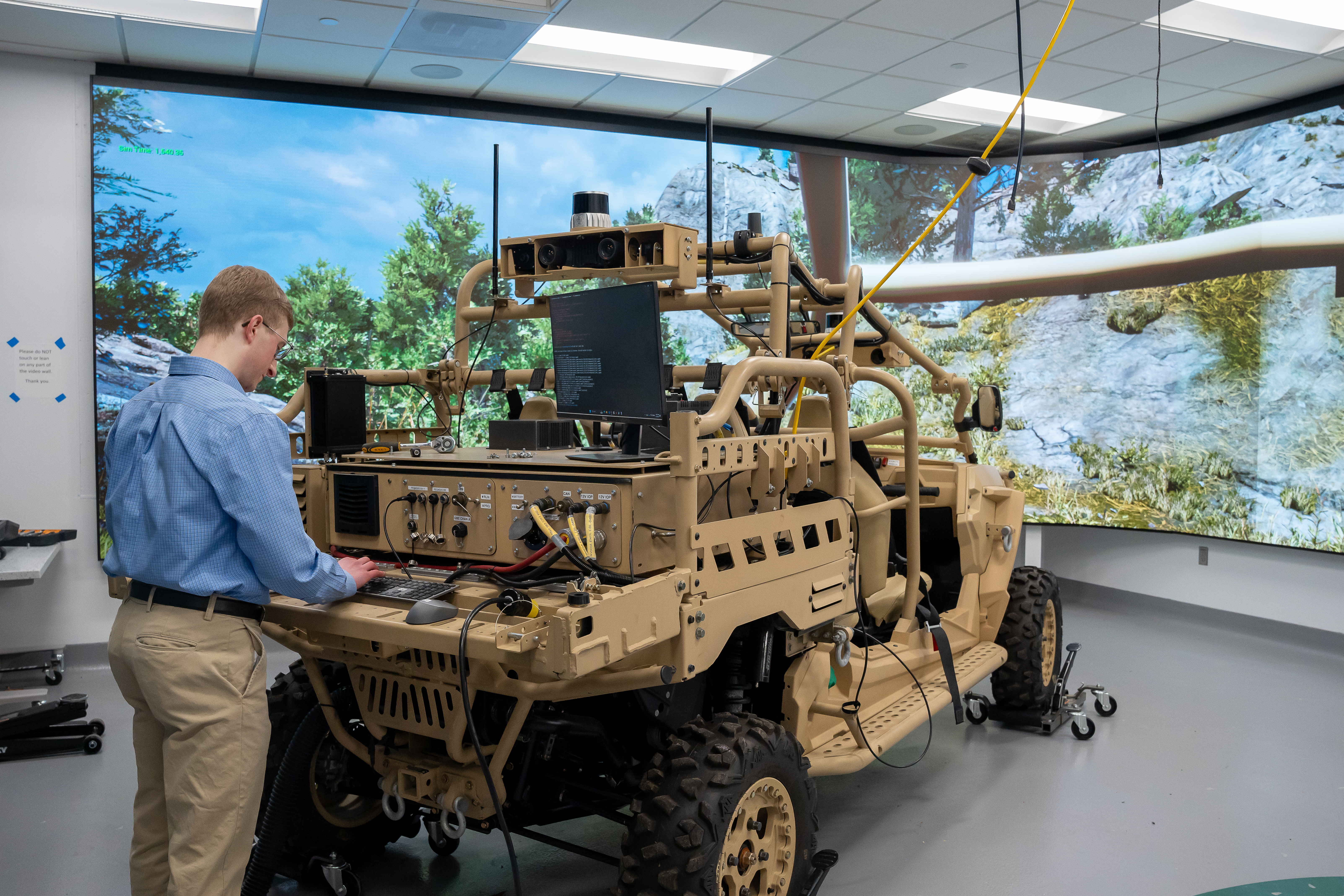Mechanical engineering PhD student Ted Sender configures the physical MRZR vehicle platform, interfaced with a synthetic off-road environment in the Digital Engineering (DE) lab of the Automotive Research Center. A wraparound screen creates an immersive experience. This DE setup supports integrated autonomy and mobility testing in mission-relevant scenarios. Photo: Levi Hutmacher/Michigan Engineering
